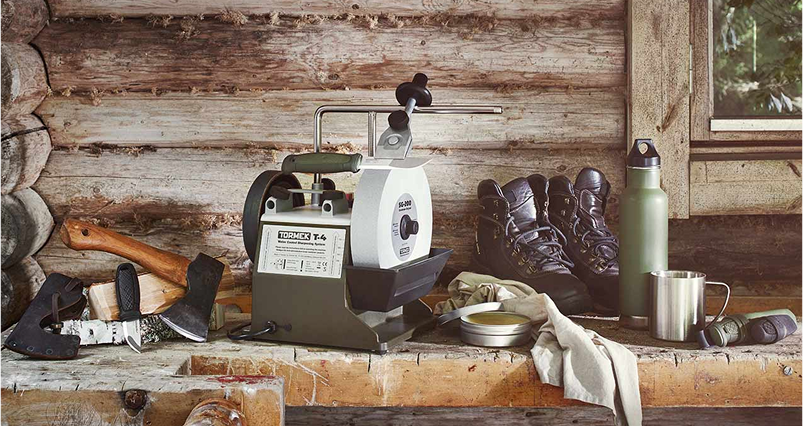 A Tormek T-4 Bushcraft sharpener on a rustic wooden table, surrounded by outdoor equipment such as axes, boots, a water bottle and a compass.