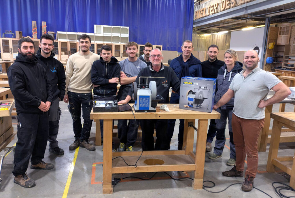Group of professionals and trainers gathered around a Tormek T-8 grinder in a carpentry workshop.