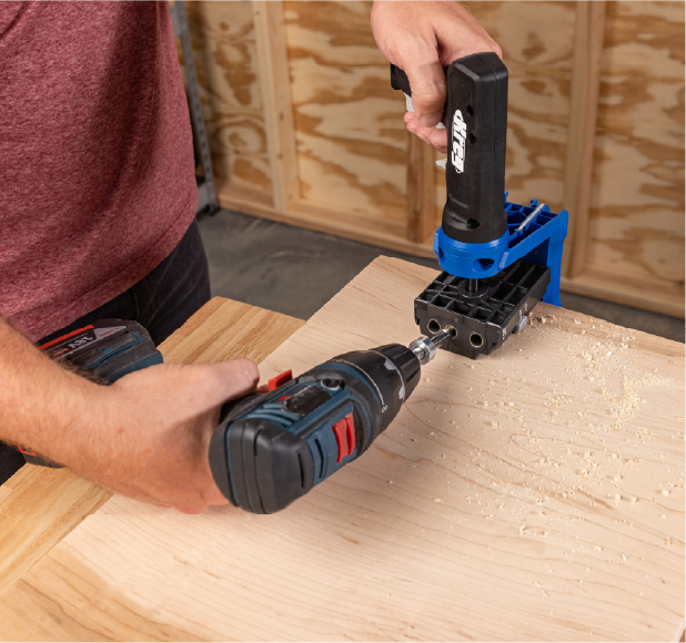 Man using a drill with a Kreg drilling jig to make angled joints on a wooden board.