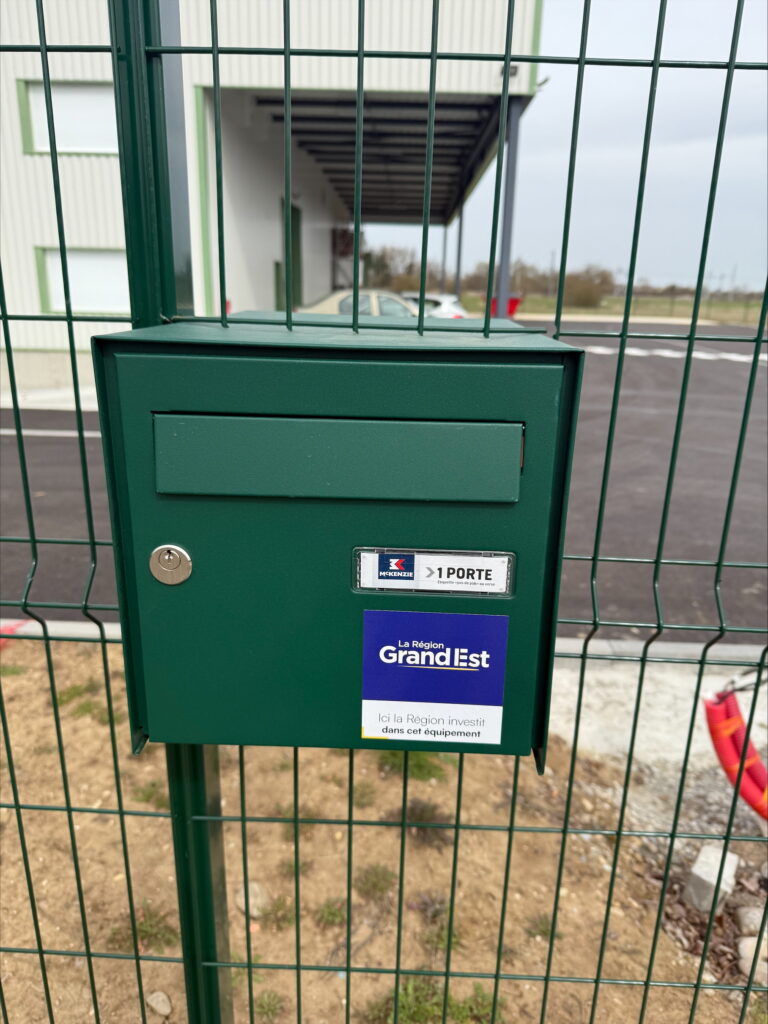 Green letterbox fixed to a fence, bearing a Région Grand Est sticker in front of the Atelier-Relais building