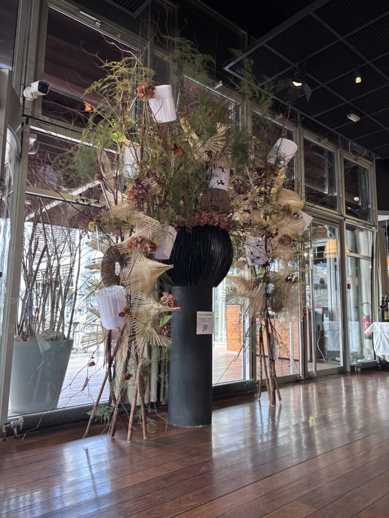 Imposing floral arrangement with branches, dried foliage and white decorative elements in a large black vase, displayed in a modern glass interior.