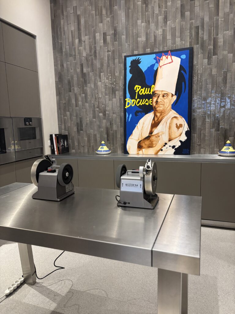 Two Tormek sharpening machines on a stainless steel table in a modern kitchen, with a colourful portrait of Paul Bocuse in the background.
