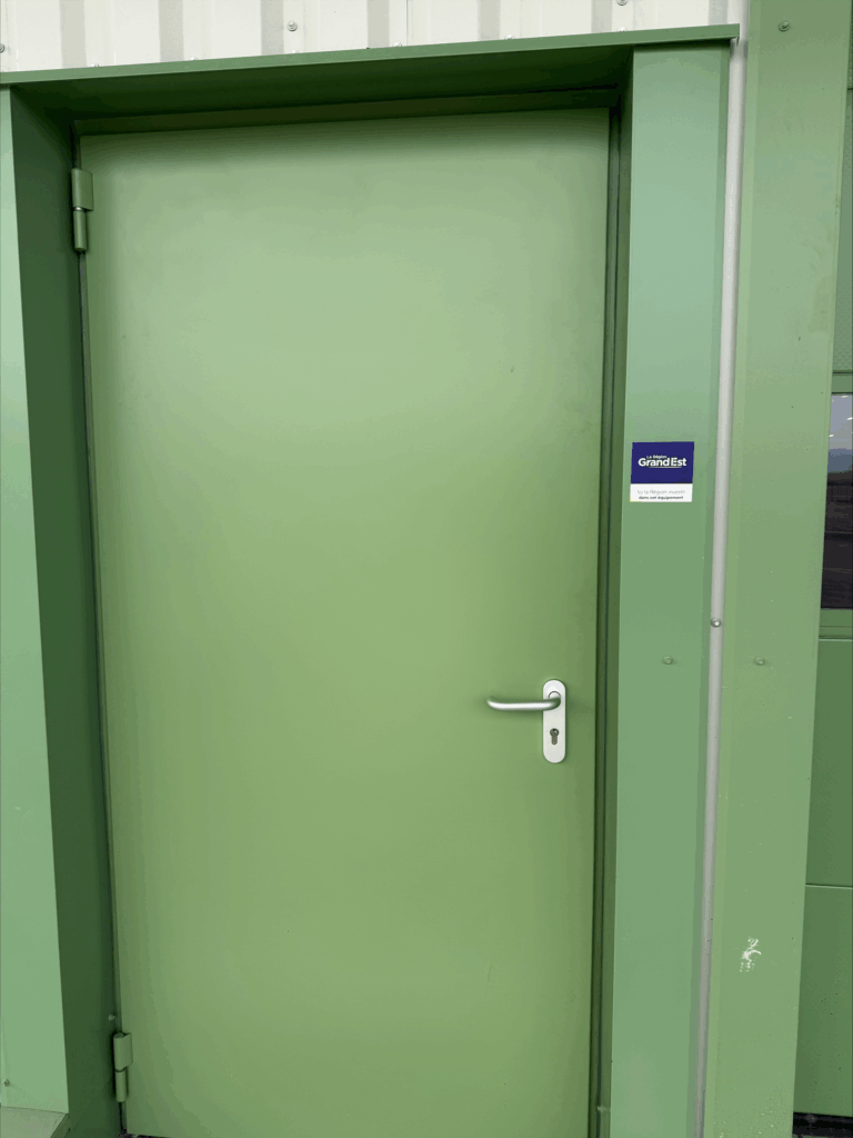 Green metal door with white handle, displaying a Région Grand Est sticker